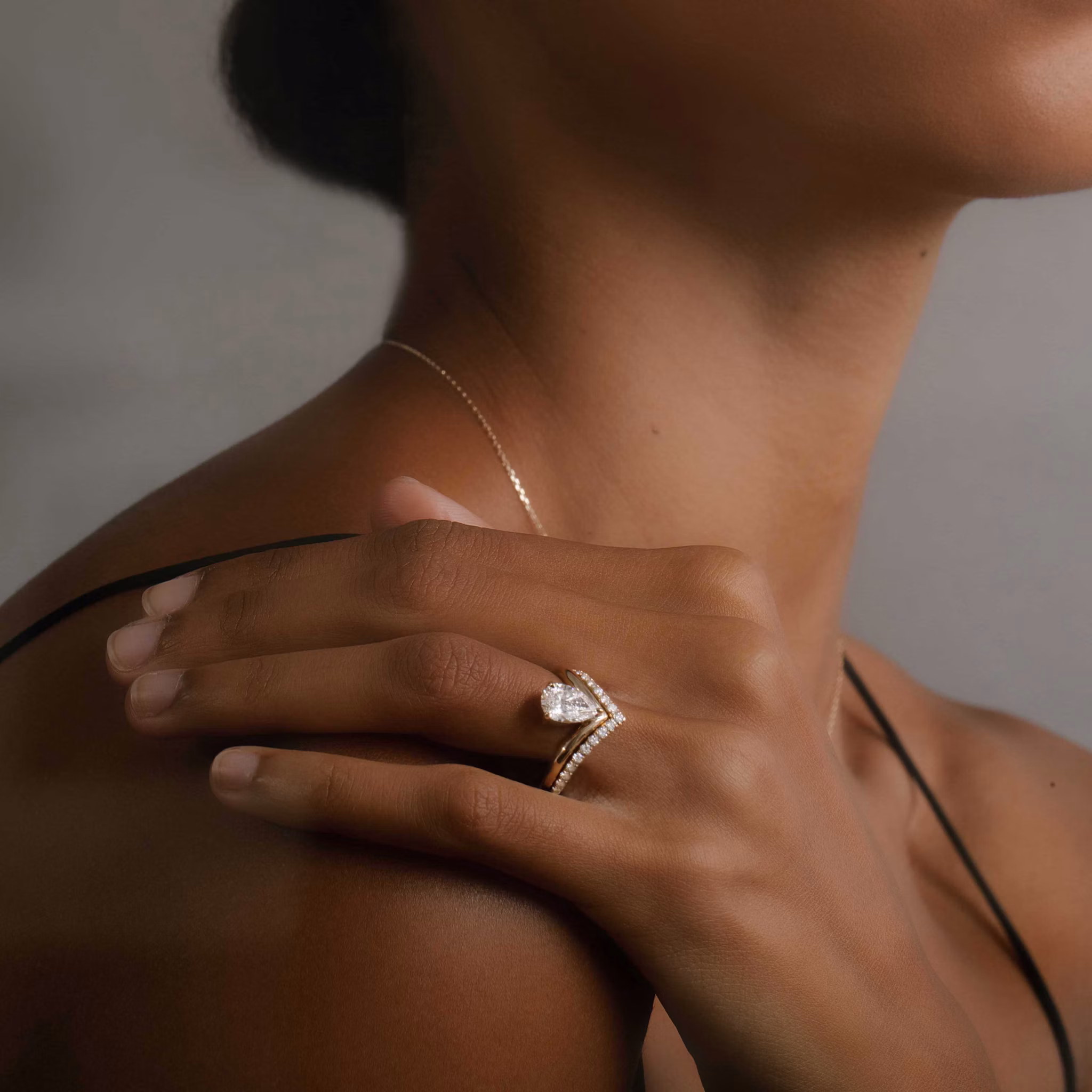 A model wears a gold engagement ring featuring a prominent pear-cut diamond paired with a matching pavé-set V-shaped band.