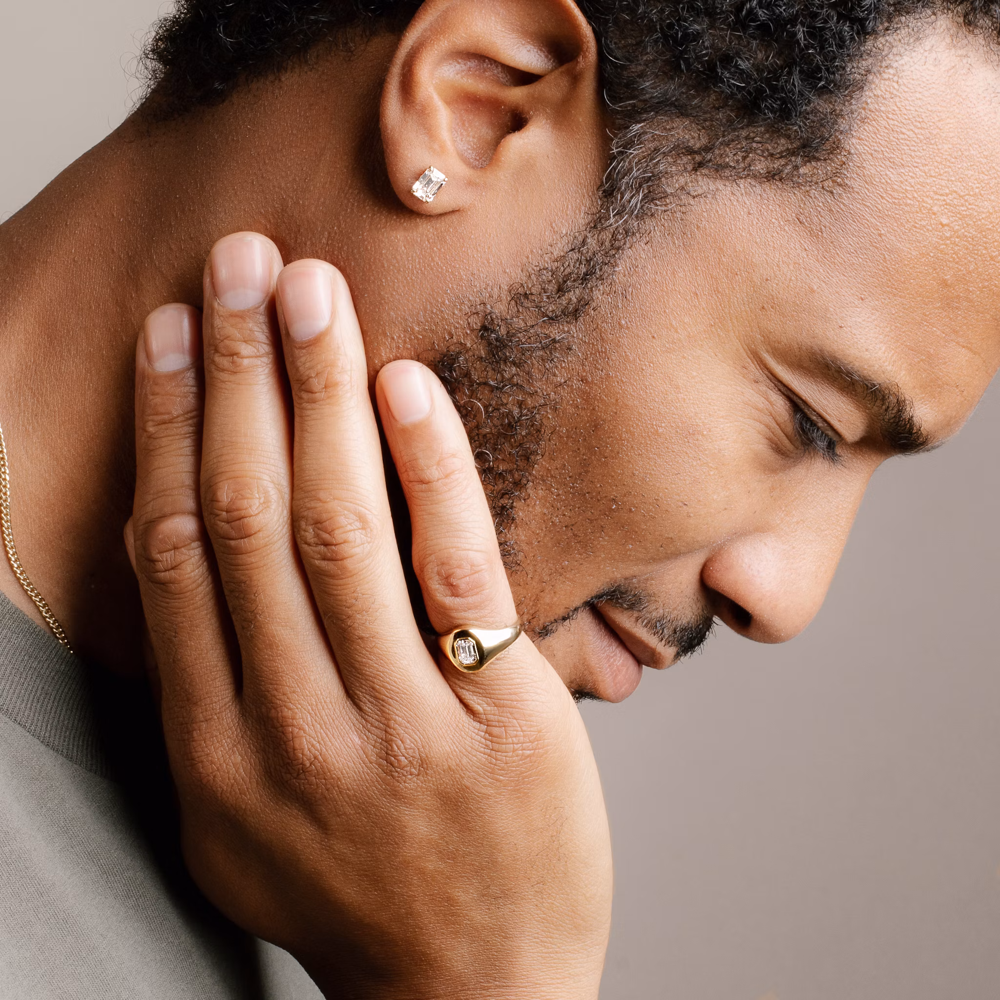 A close-up profile of a man with short hair and a beard, wearing an emerald-cut diamond stud earring and a gold signet ring featuring a matching emerald-cut diamond.