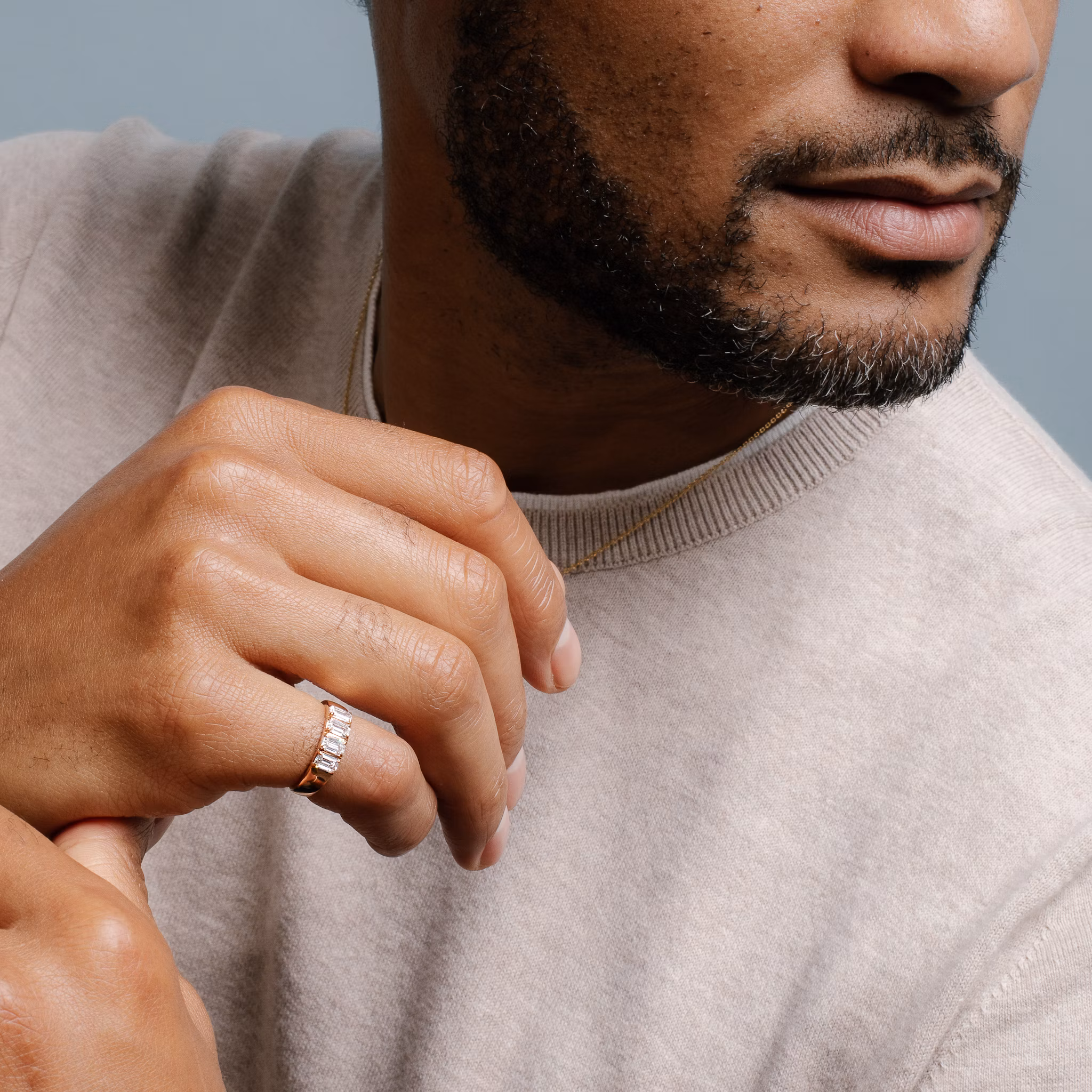 Close-up of a man's manicured hand touching his shoulder, showcasing a gold ring with four emerald-cut diamonds on his pinky finger.