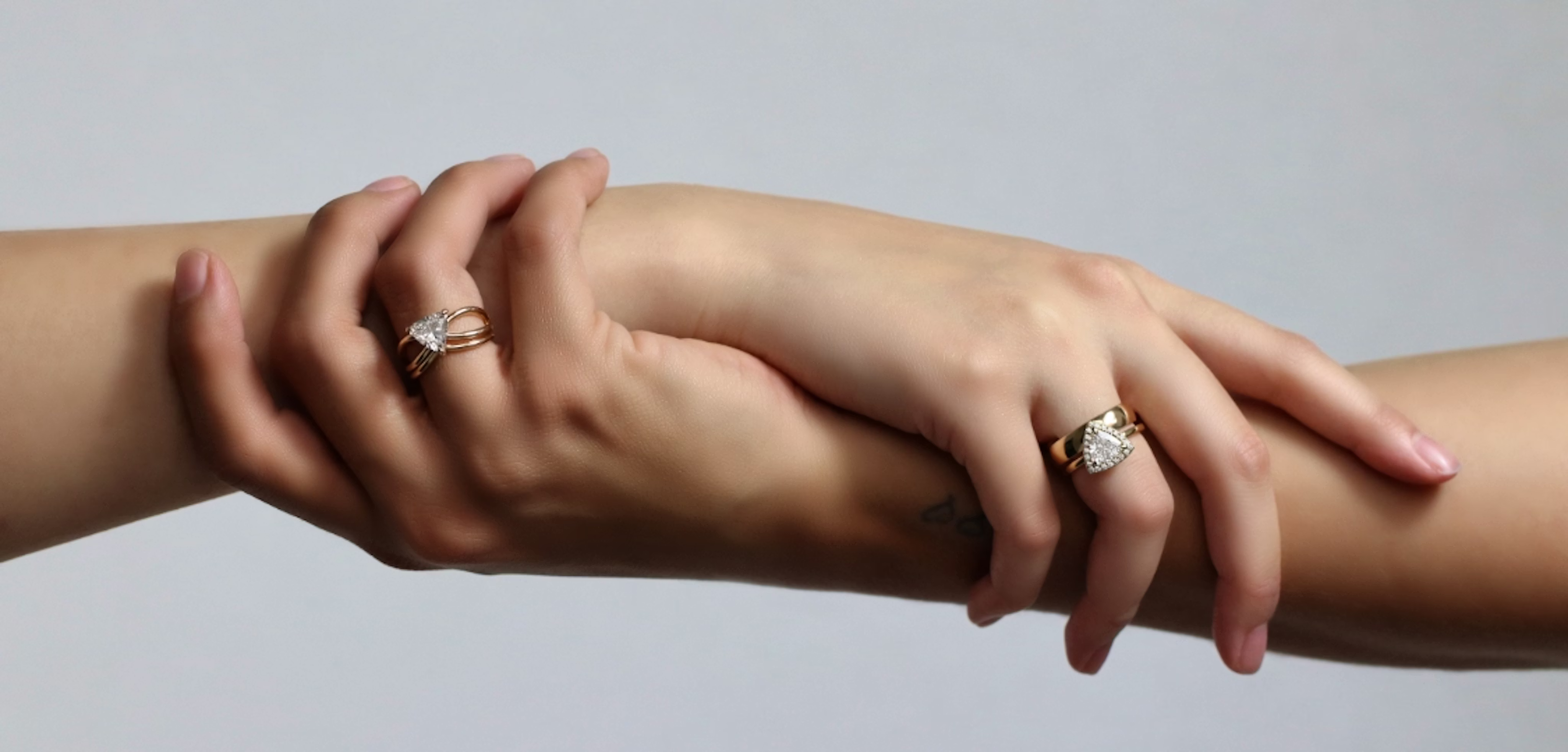 A close-up image of two hands reaching across the frame, each wearing a diamond engagement ring.