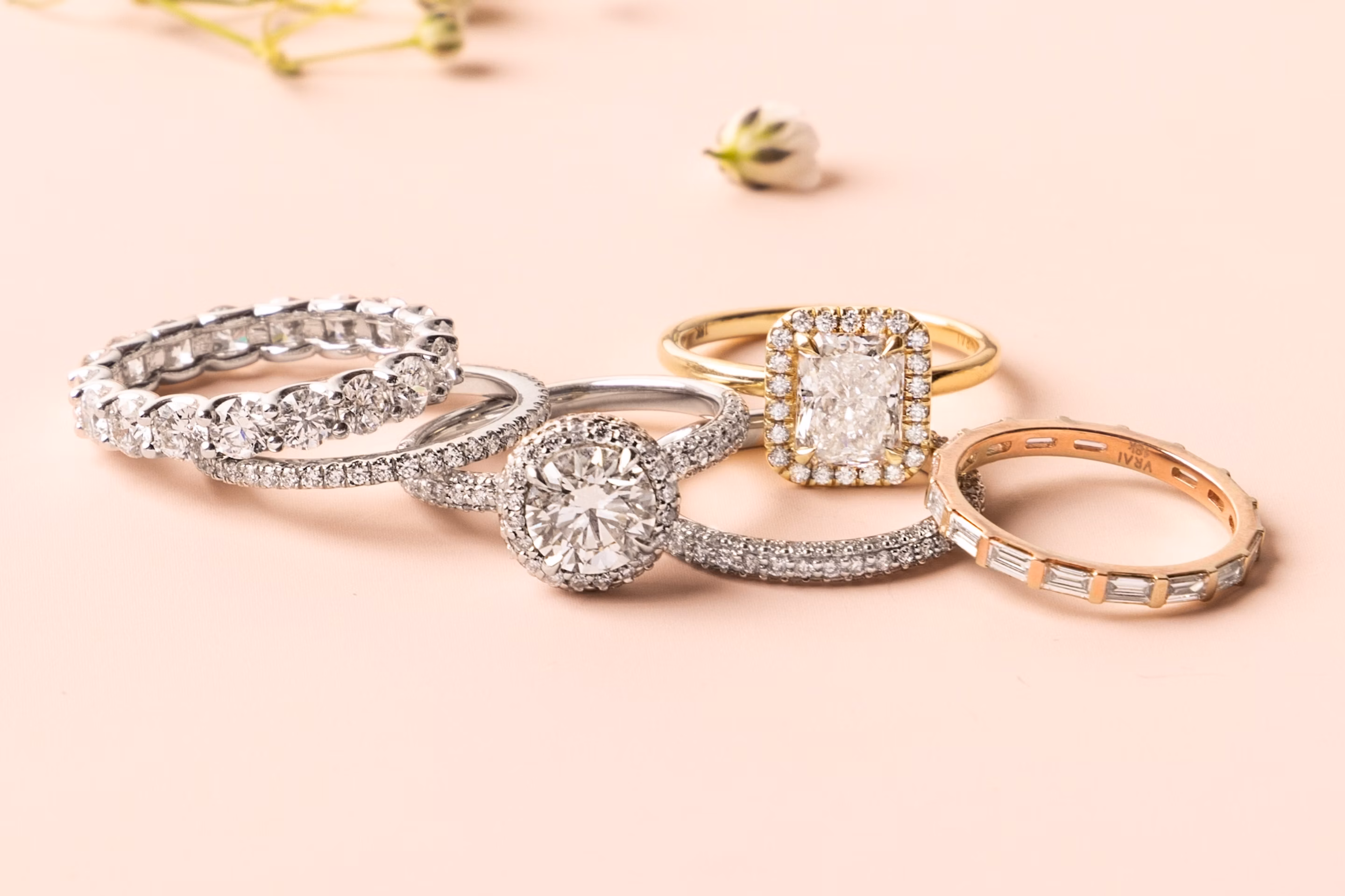 Five diamond rings in platinum, yellow gold, and rose gold, featuring round, oval, and emerald-cut stones on a blush background.