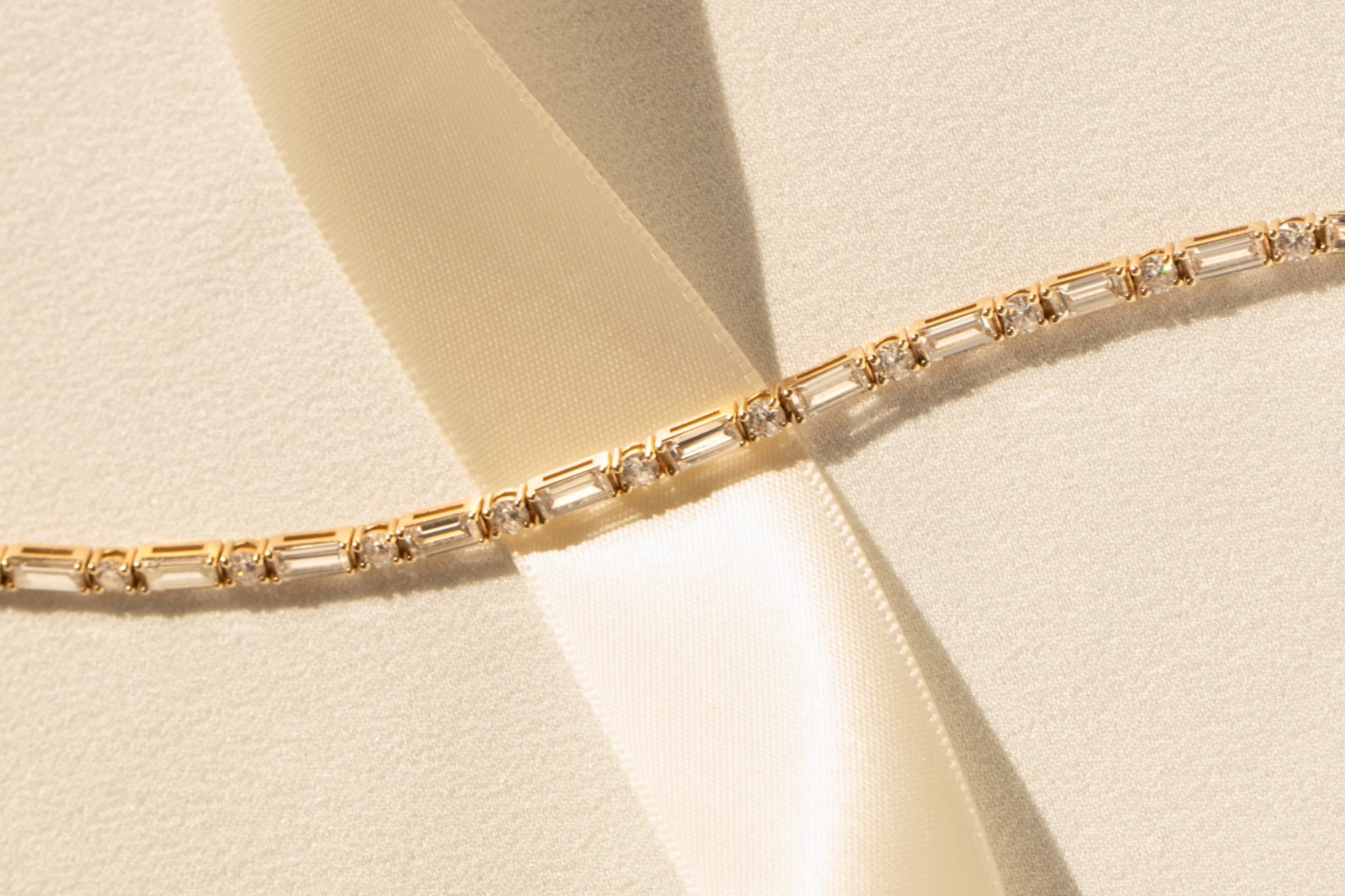 A close-up shot of a yellow gold tennis bracelet resting diagonally across a soft cream-colored fabric ribbon.
