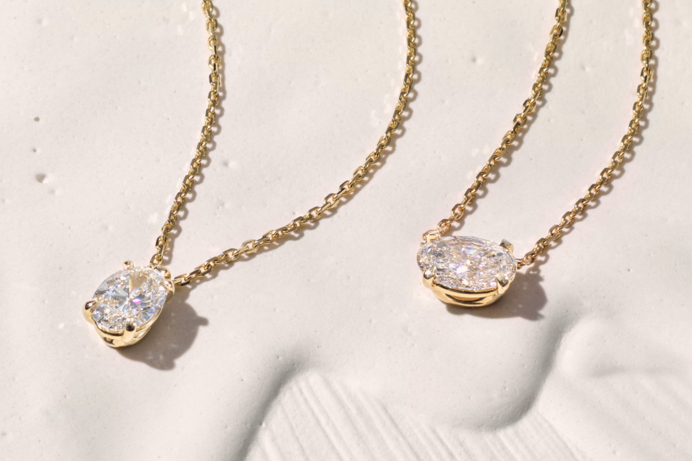 Two gold chain necklaces with oval-cut diamond pendants displayed on a textured white surface in soft lighting.