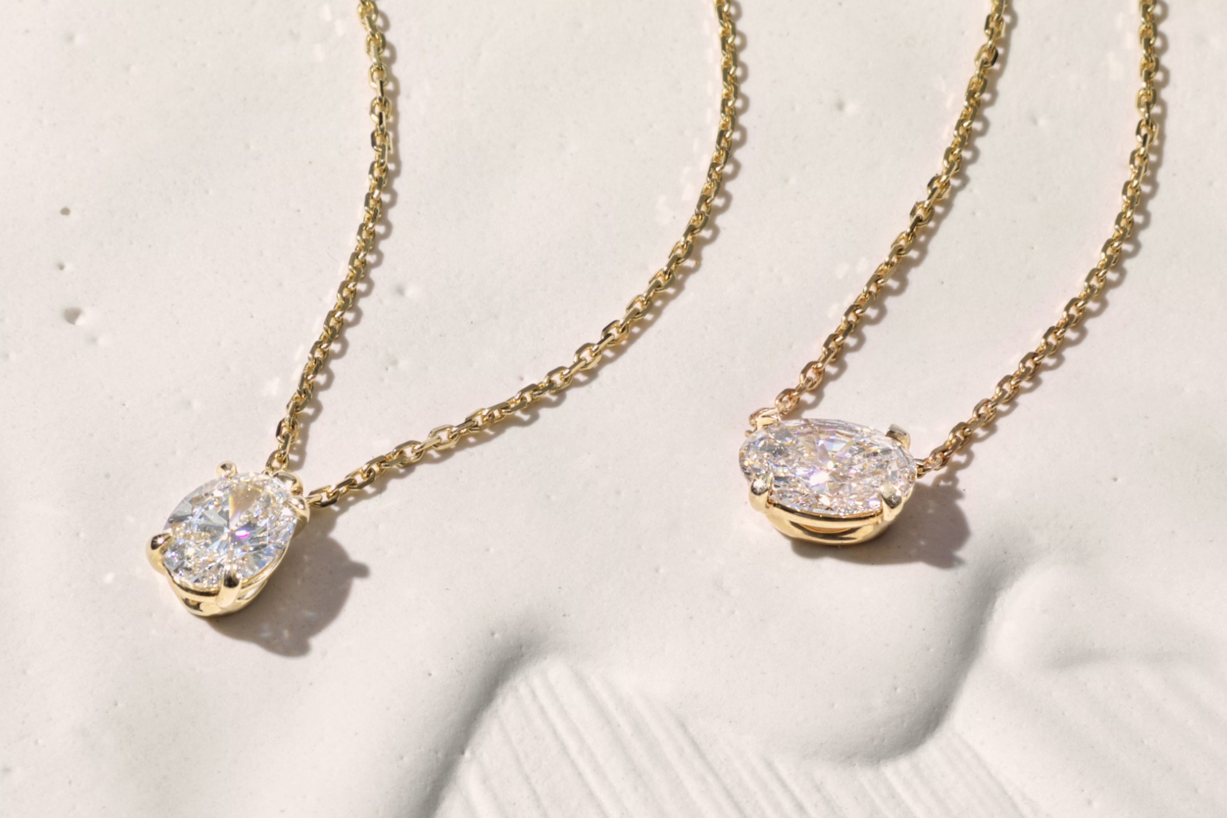 Two gold chain necklaces with oval-cut diamond pendants displayed on a textured white surface in soft lighting.