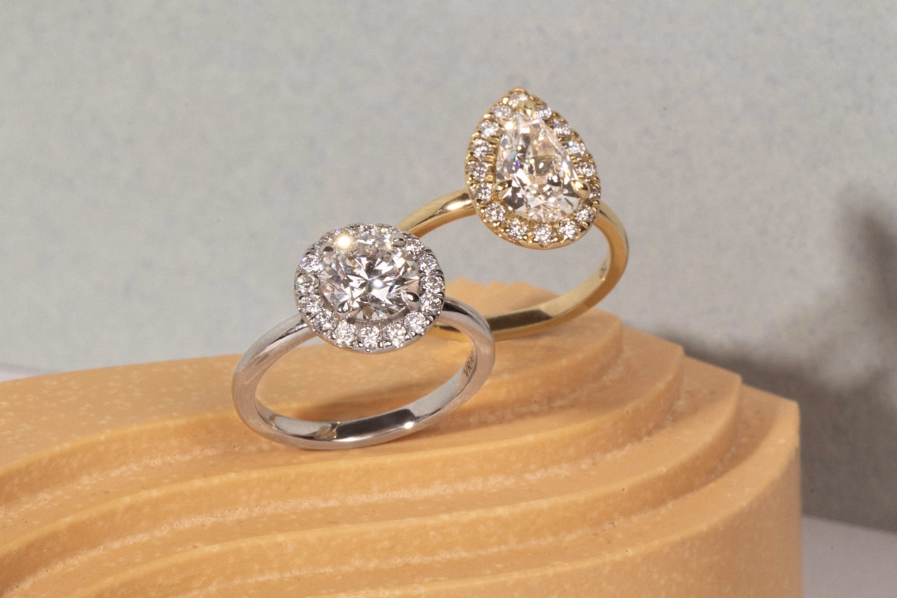 Two diamond halo cocktail rings—one with a round center stone in white gold and one pear-shaped in yellow gold—on a sculpted display