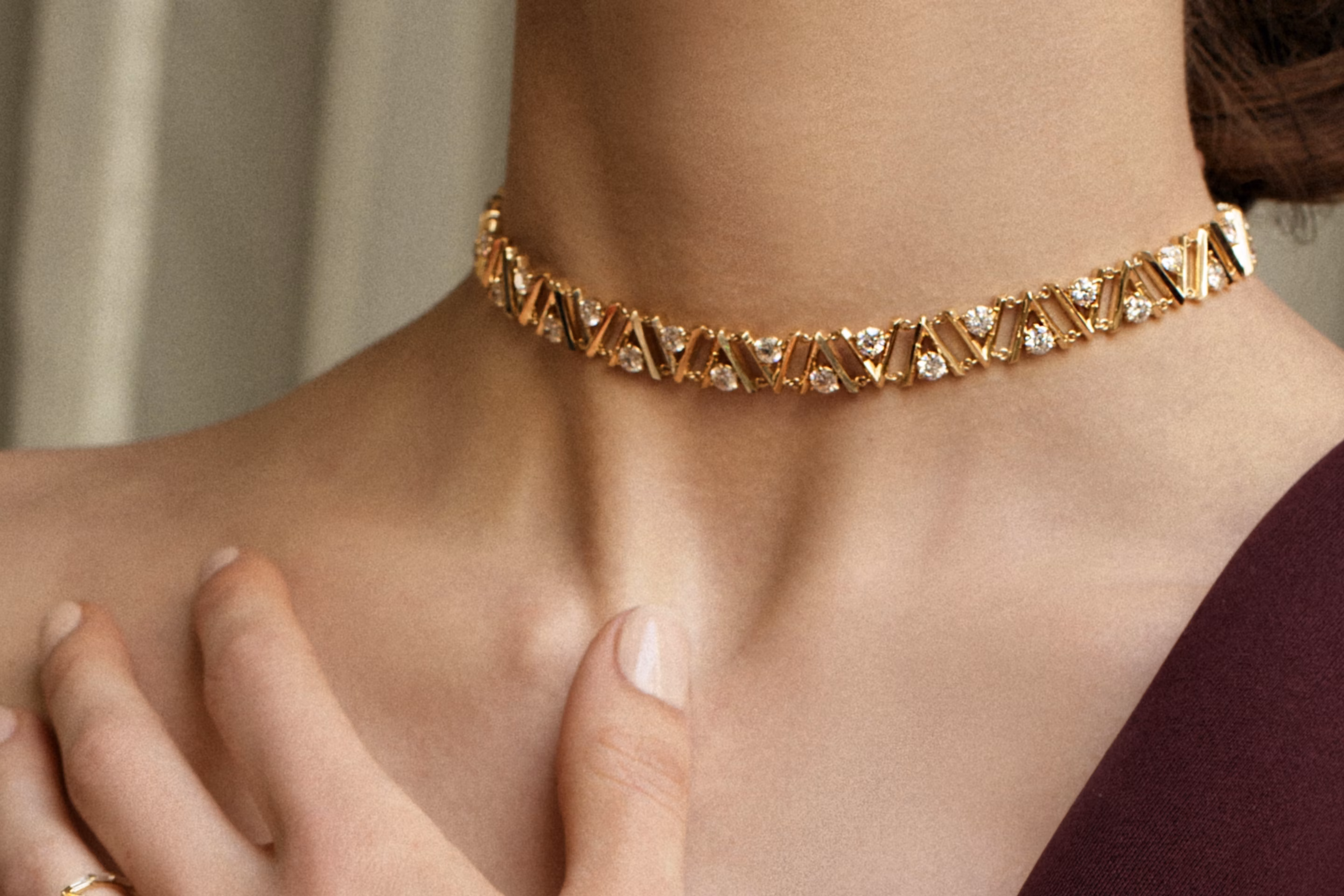 Close-up of a woman's neck wearing a gold choker featuring repeating V-shaped links and bezel-set round diamonds, paired with a burgundy one-shoulder dress.