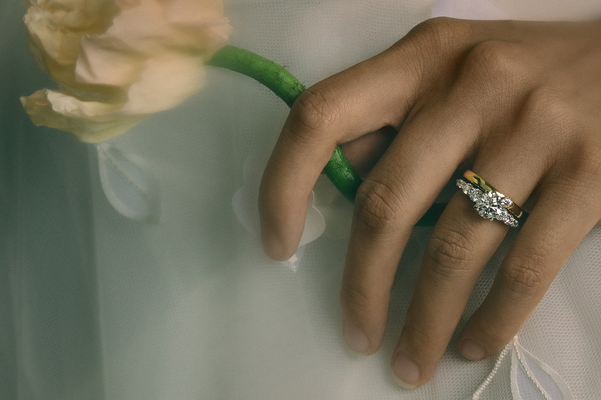 person wearing white holding a flower with engagement ring in platinum or white gold and wedding band in yellow gold visible