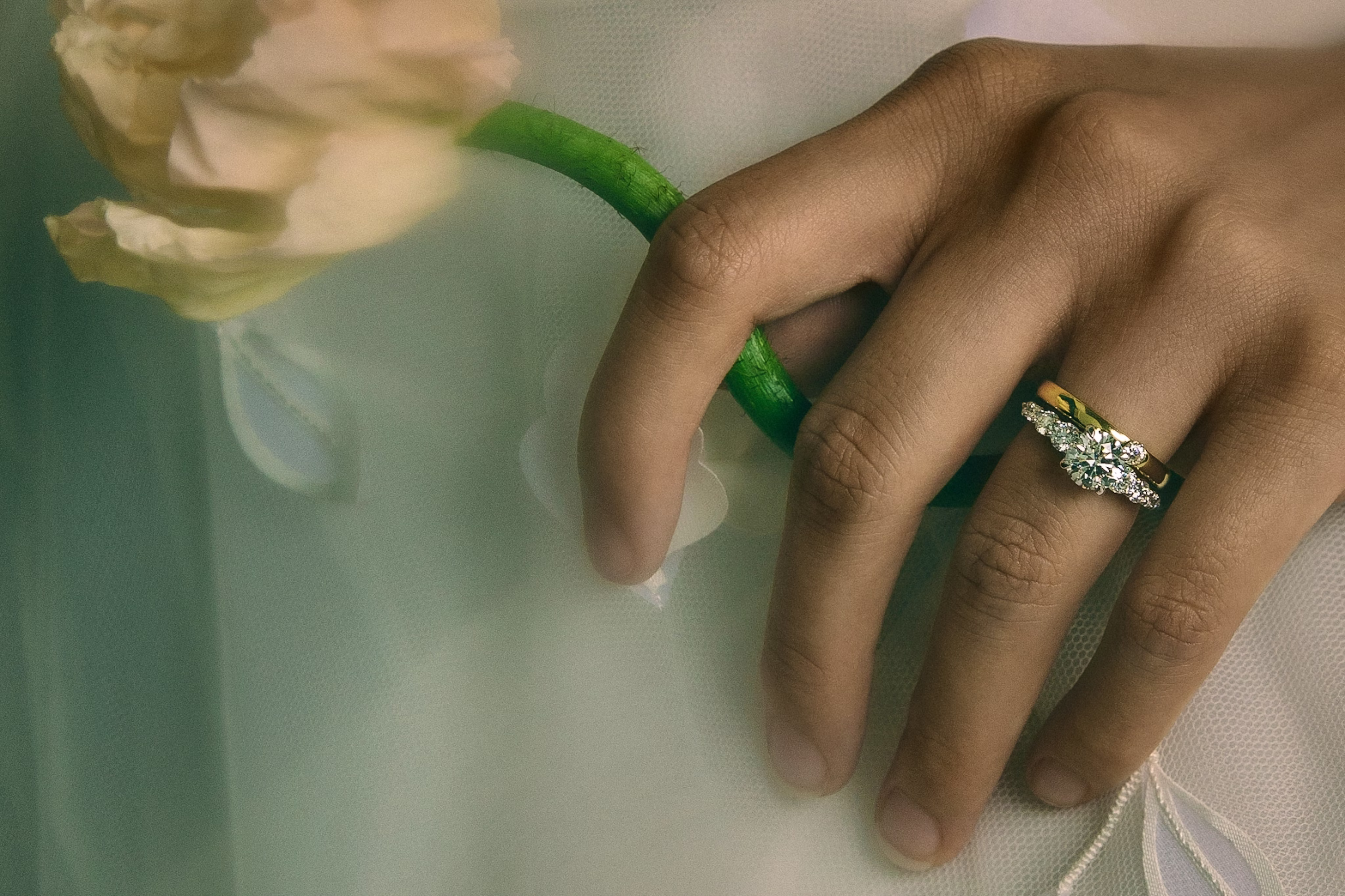 person wearing white holding a flower with engagement ring in platinum or white gold and wedding band in yellow gold visible