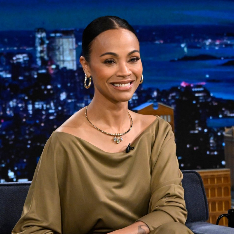 An image of Zoe Saldana wearing our Oval Dome Hoops, Collar with Drop Pendant, and Dome Cuff Bracelet all in gold, while sitting in a chair on the set of The Tonight Show starring Jimmy Fallon.