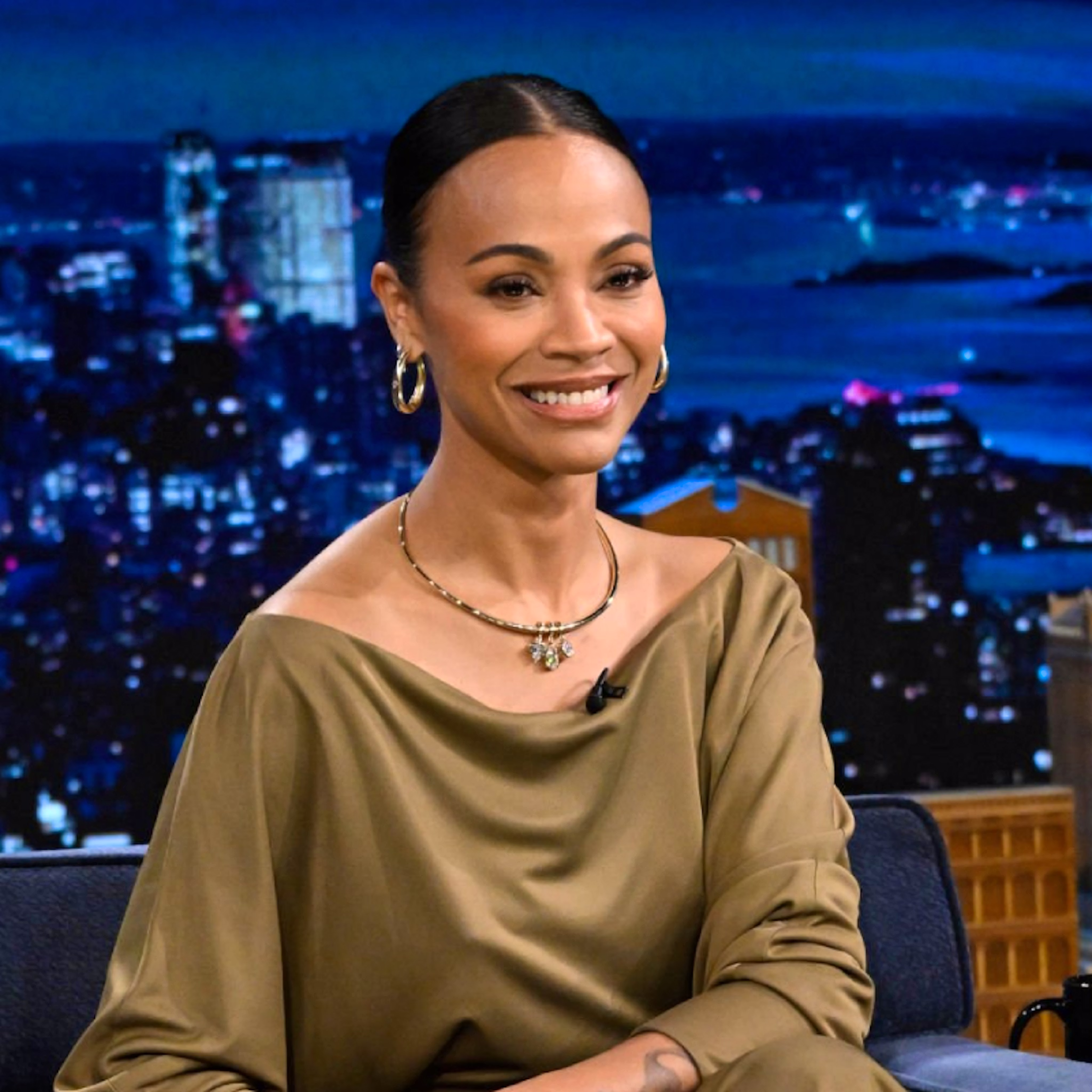 An image of Zoe Saldana wearing our Oval Dome Hoops, Collar with Drop Pendant, and Dome Cuff Bracelet all in gold, while sitting in a chair on the set of The Tonight Show starring Jimmy Fallon.