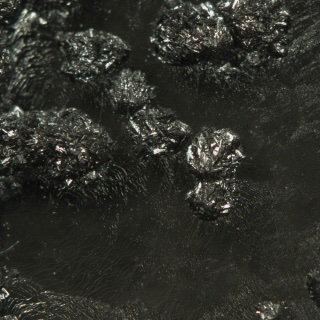 A close-up photograph of a dark mineral, showing bumpy, rounded clumps with many shiny, reflective facets.