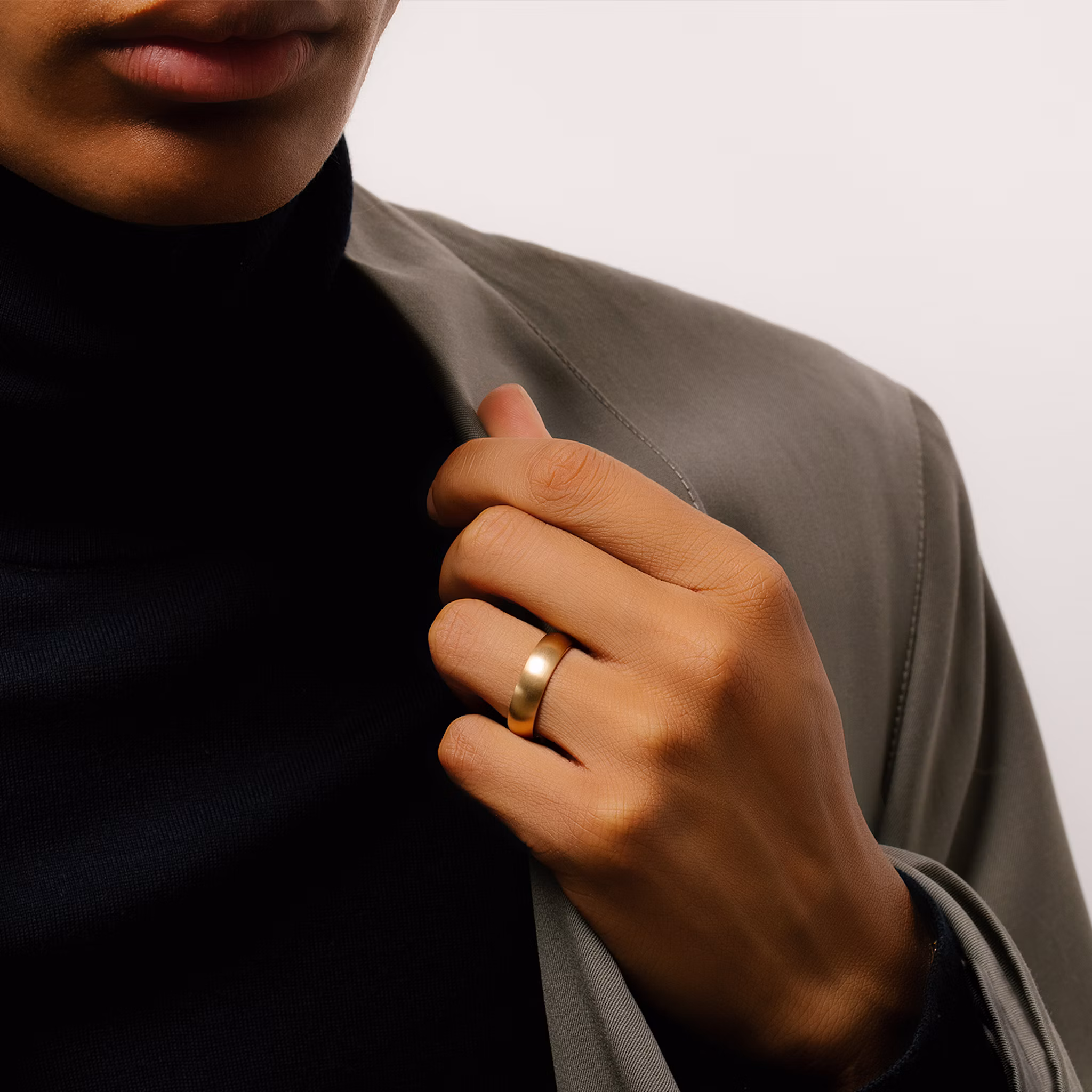 Close-up of a person wearing a gold ring on their ring finger, with their hand resting on the lapel of a gray blazer over a black turtleneck.