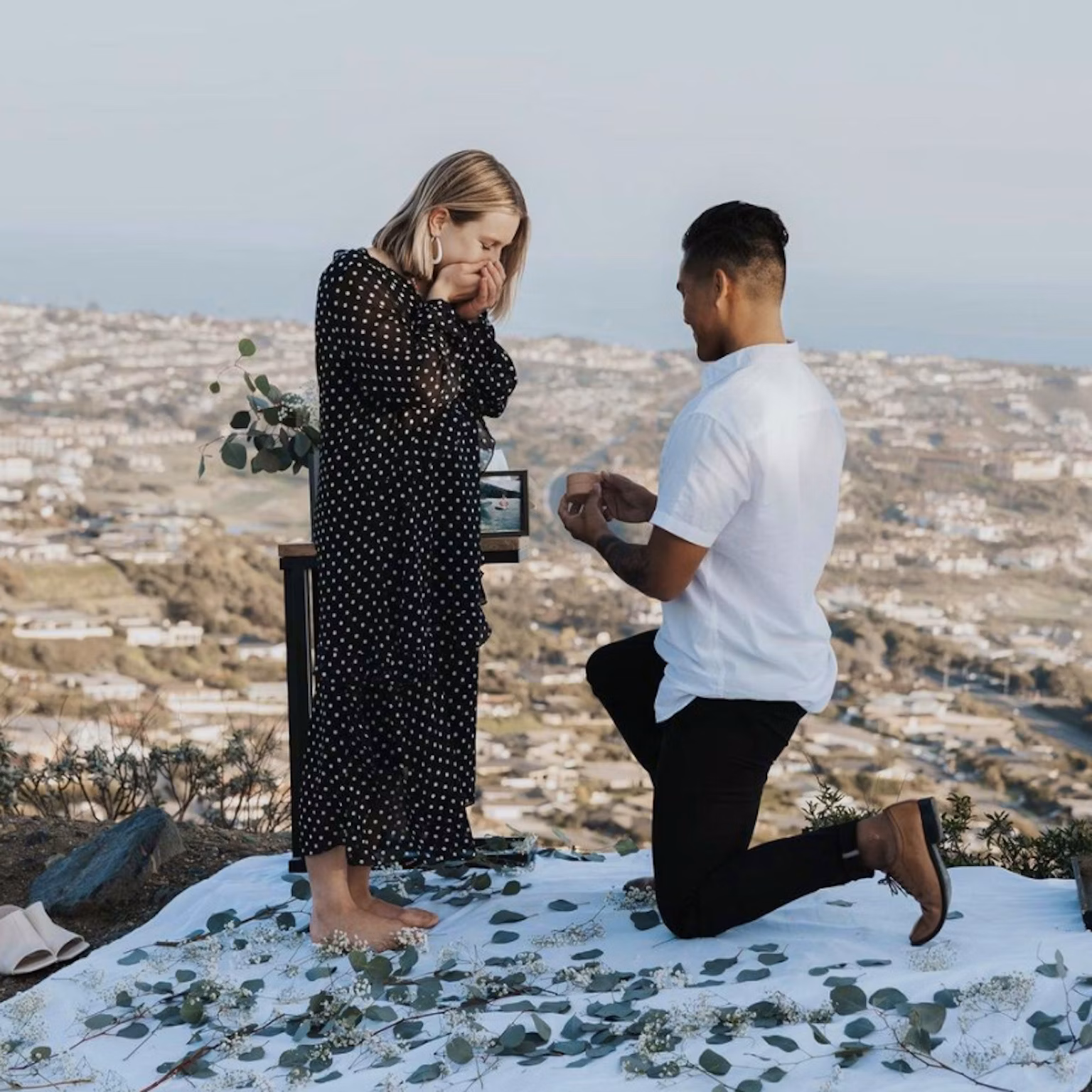 A person is proposing to their partner, on a white picnic blanket that is scattered with greenery.