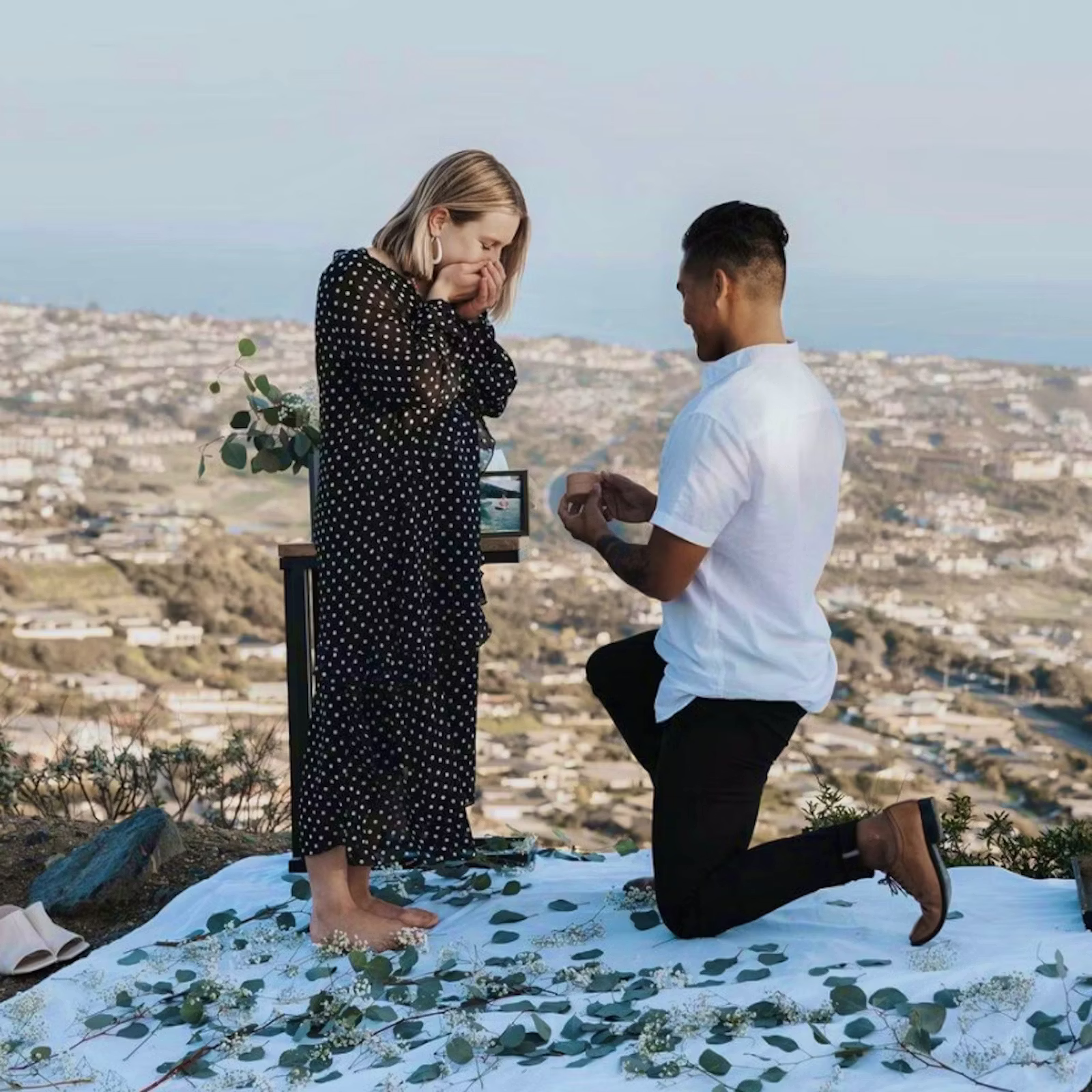 A person is proposing to their partner, on a white picnic blanket that is scattered with greenery.