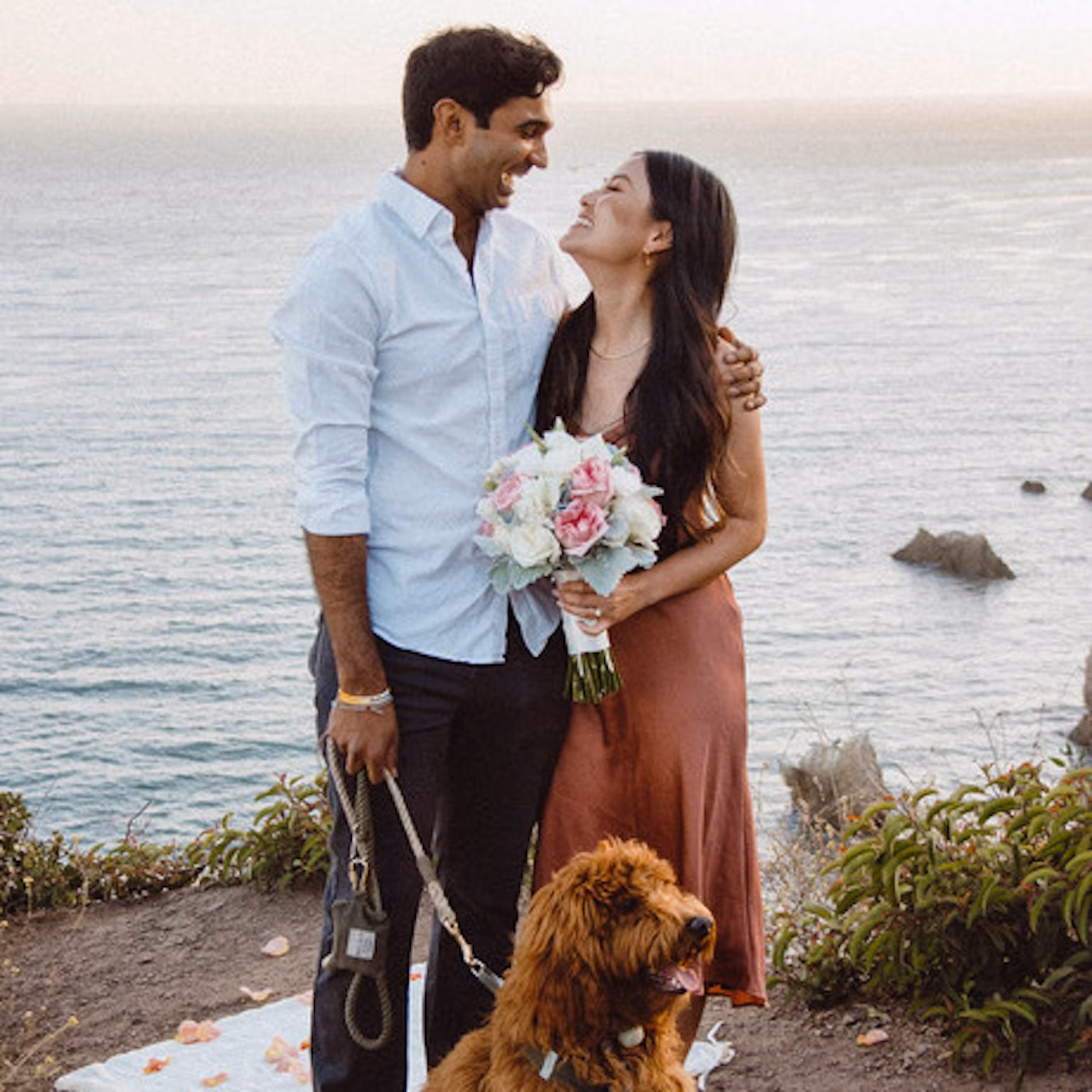 A couple looking at each other, with the woman holding a bouquet and the man holding a leash attached to a dog.
