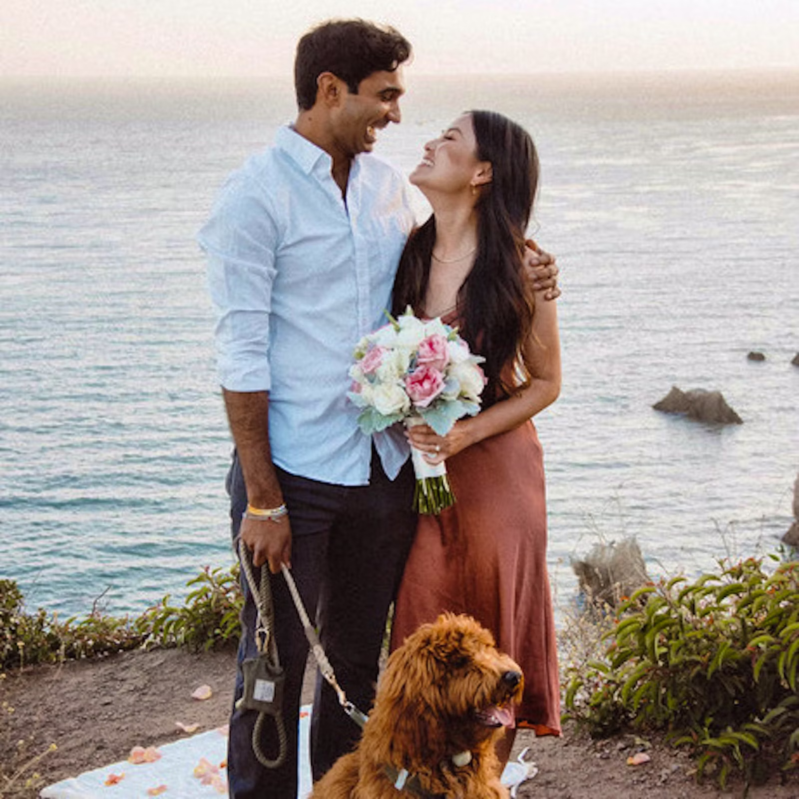 A couple looking at each other, with the woman holding a bouquet and the man holding a leash attached to a dog.