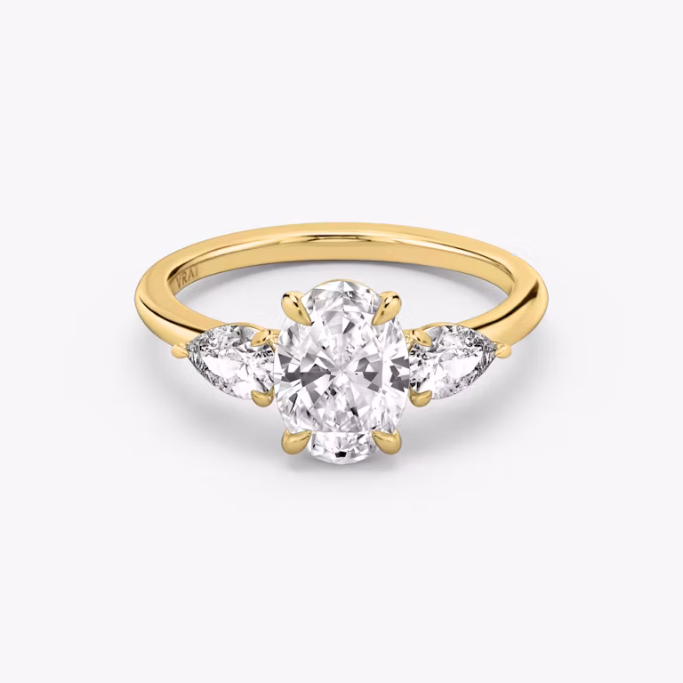 A close-up image of VRAI's Three Stone Oval Yellow Gold Engagement Ring, lying flat.