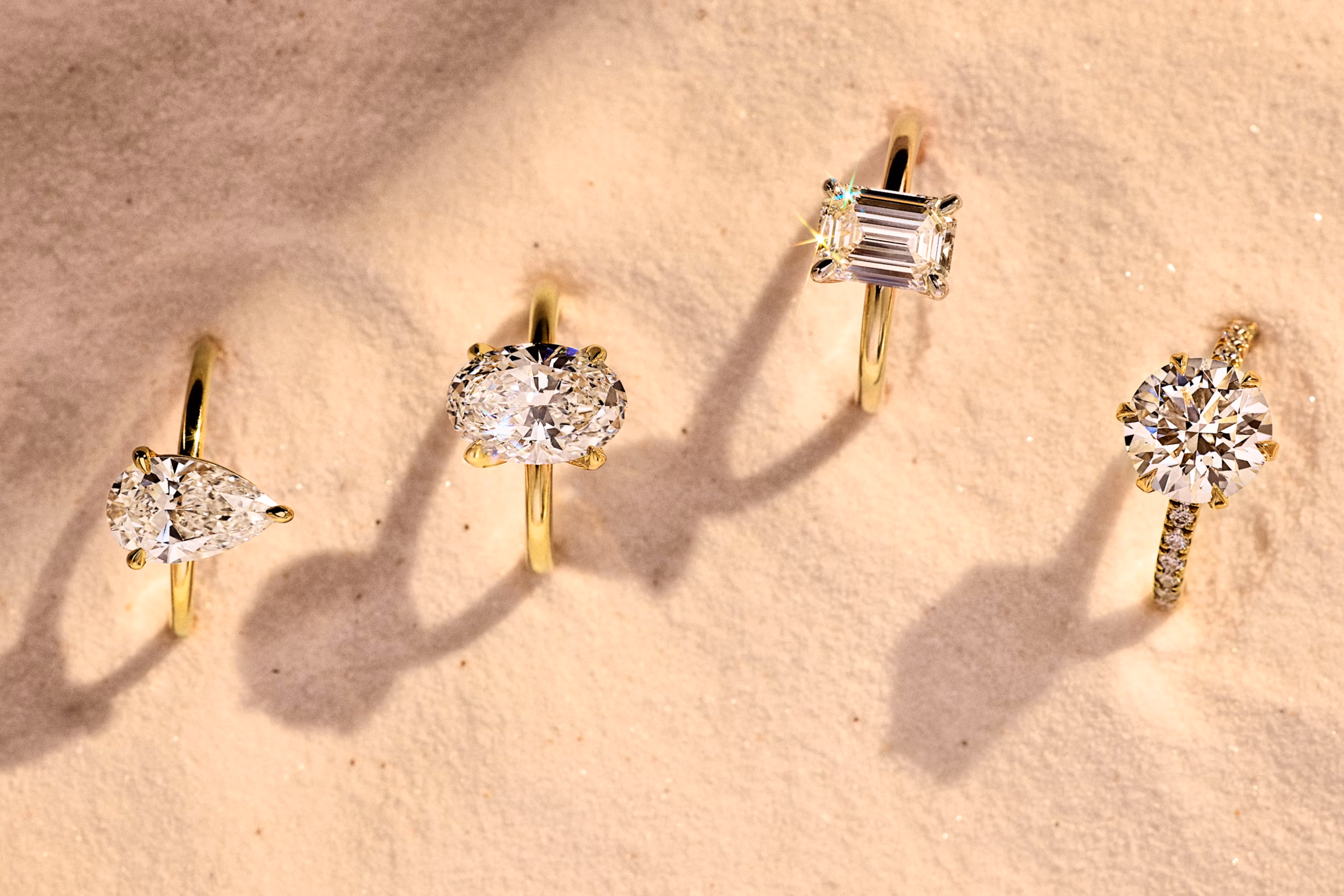 Four gold engagement rings with pear, oval, emerald, and round-cut diamonds displayed on soft sand in natural light.