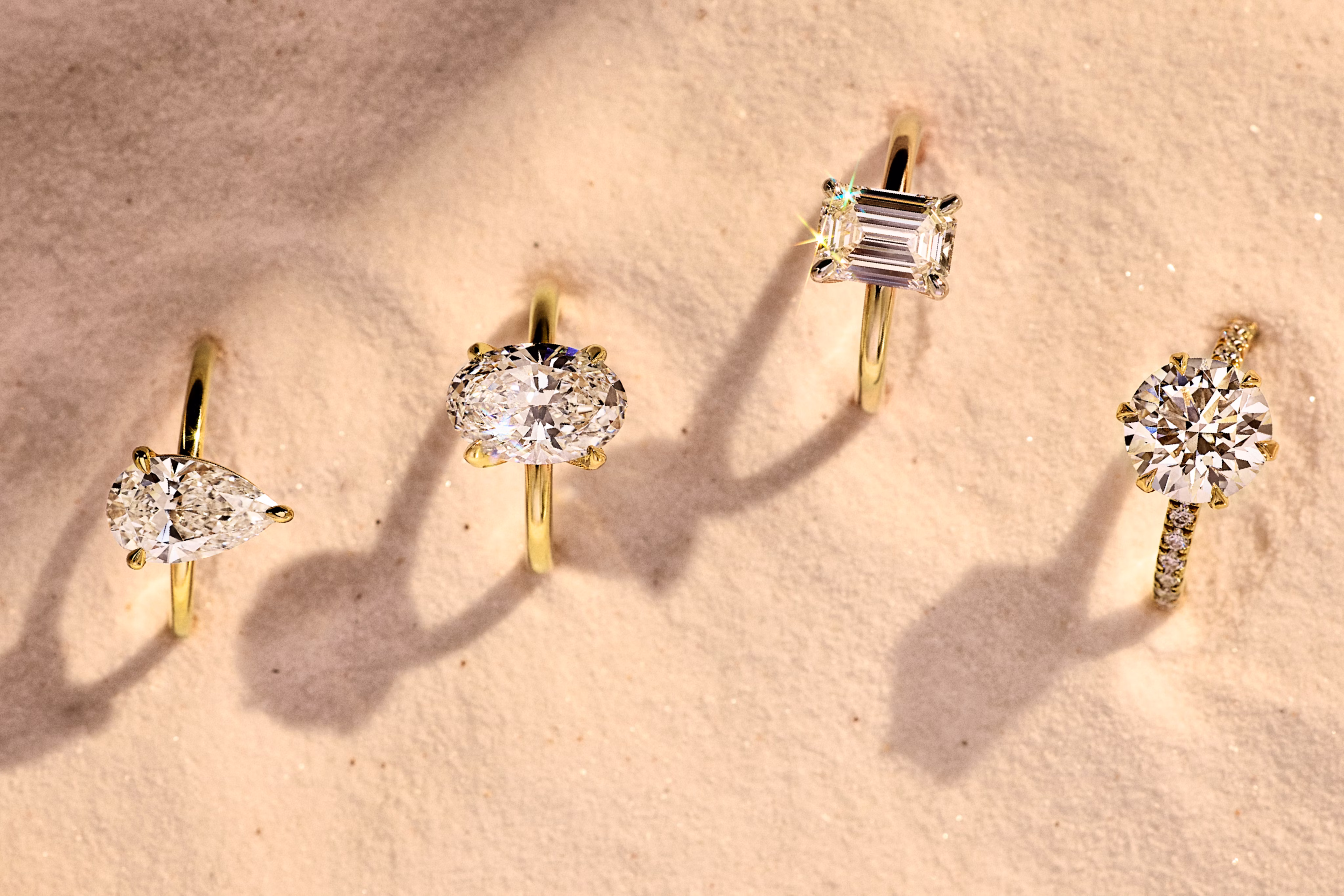 Four gold engagement rings with pear, oval, emerald, and round-cut diamonds displayed on soft sand in natural light.