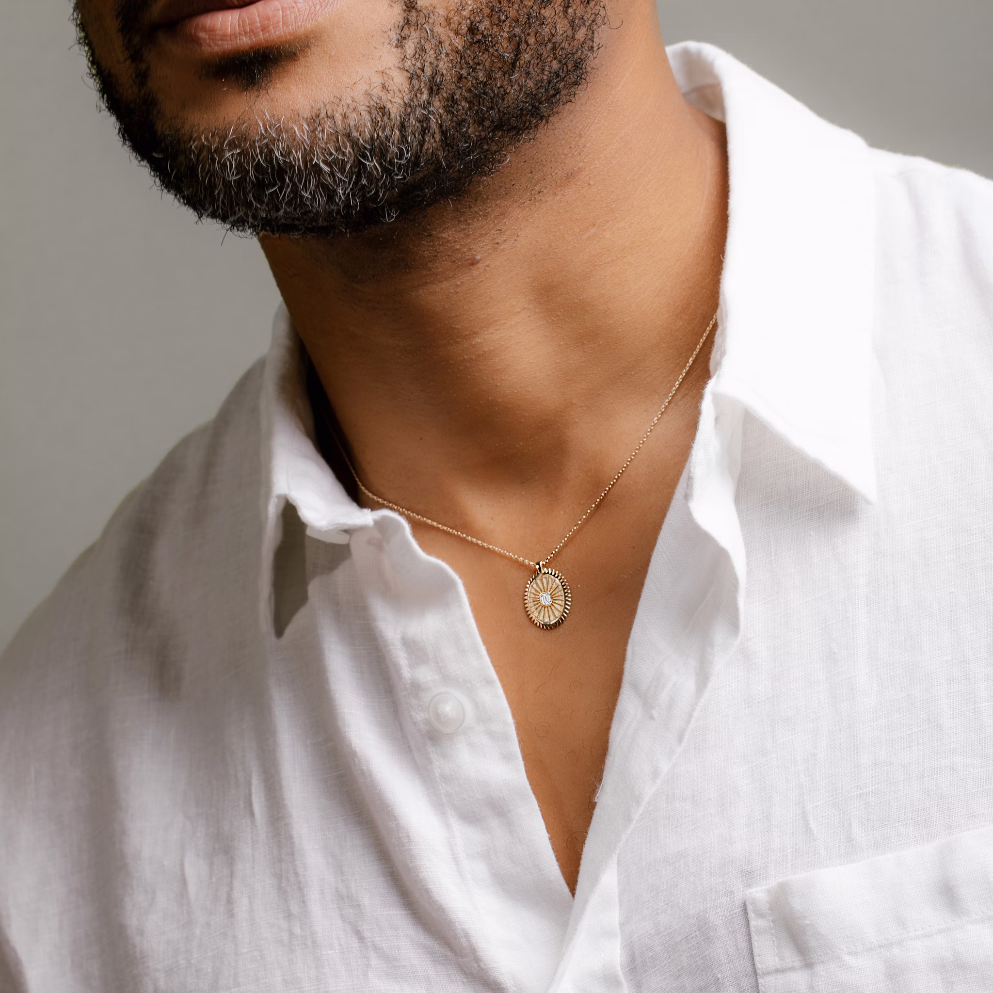Man wearing a white shirt and a yellow gold medallion necklace with an emerald cut diamond, set against a grey background.