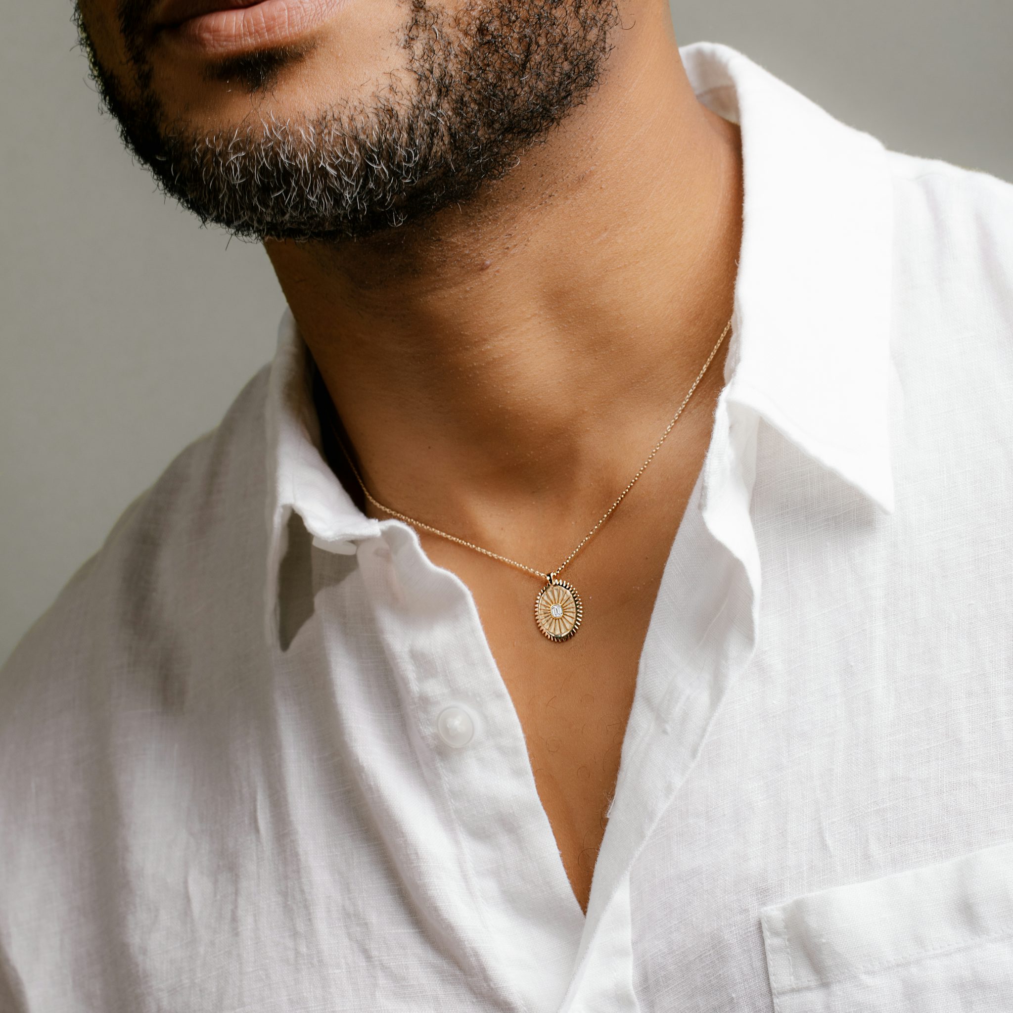 Man wearing a white shirt and a yellow gold medallion necklace with an emerald cut diamond, set against a grey background.